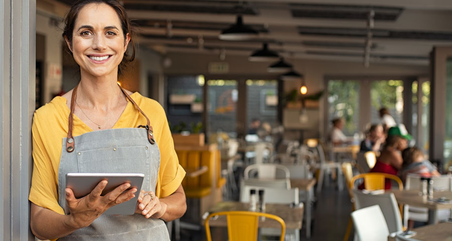 Portrait of a happy woman standing outside her store. Cheerful, mature waitress waiting for customers in the cafeteria. Successful small business owner with casual gray apron at the entrance.